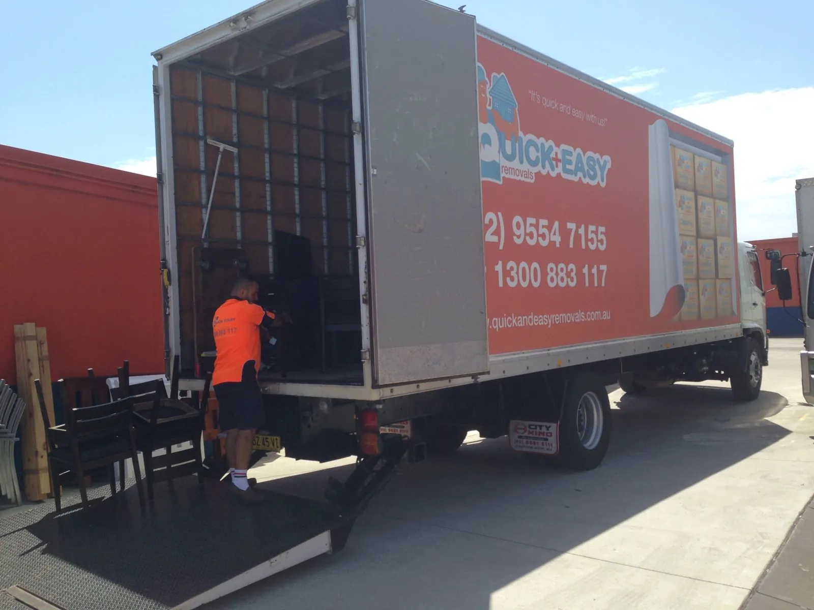 Removalist loading a dark item into a branded orange Quick and Easy truck from the tail lift