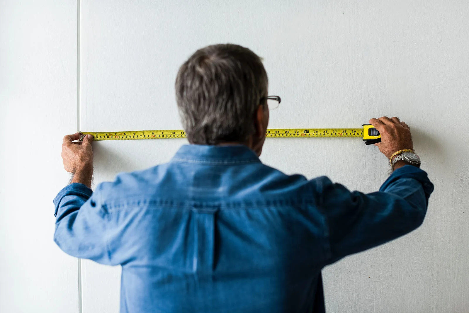A man in a denim shirt, seen from behind, stretching a yellow tape measure across a plain white wall before a move