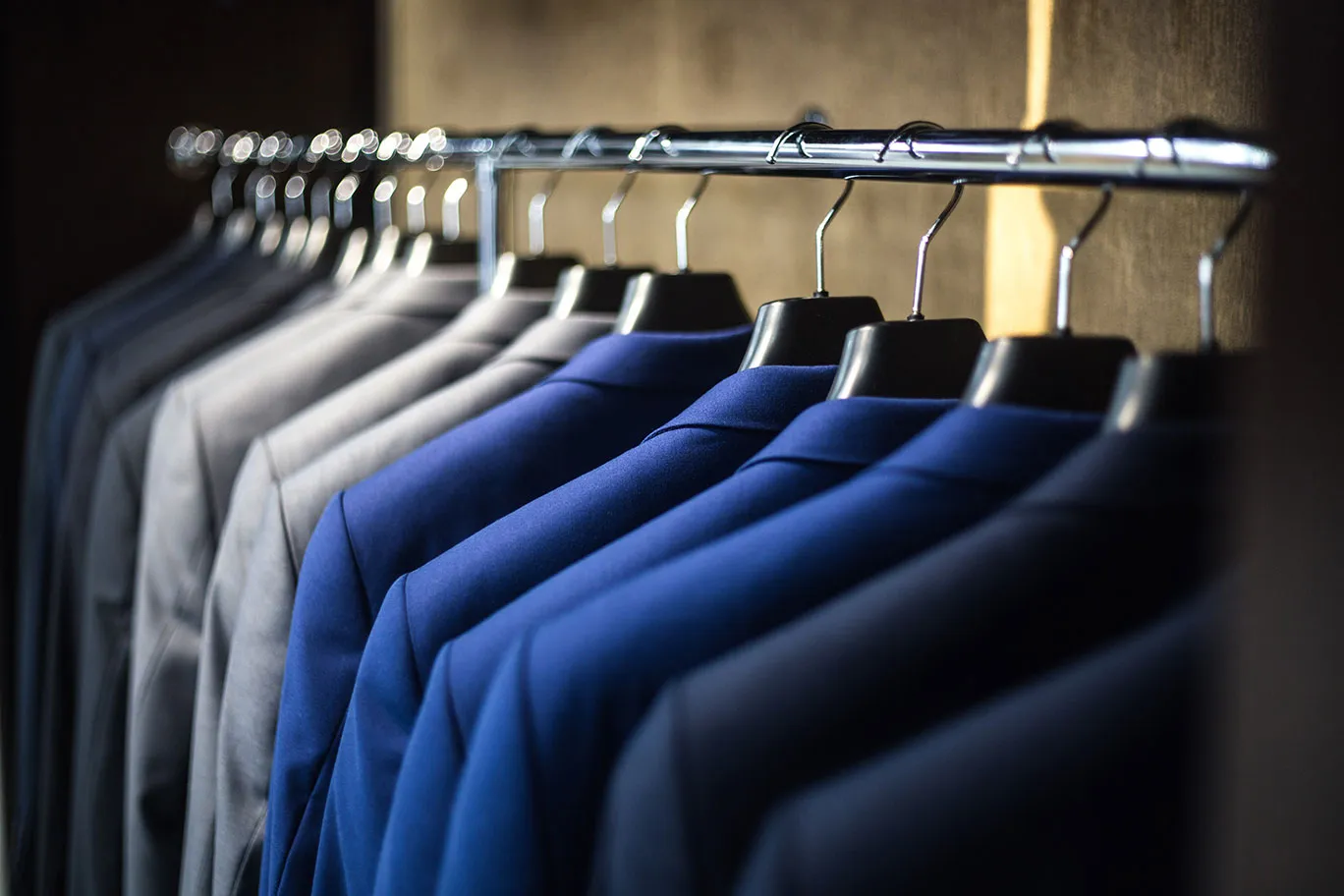 A row of blue, grey and navy suit jackets on black hangers along a chrome rail inside a dark wardrobe