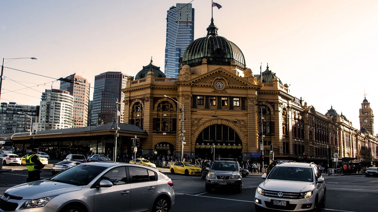 Flinders Street Station in Melbourne at dusk, its domed sandstone facade and clock lit by low sun, with cars and taxis crossing the intersection in front and city towers behind