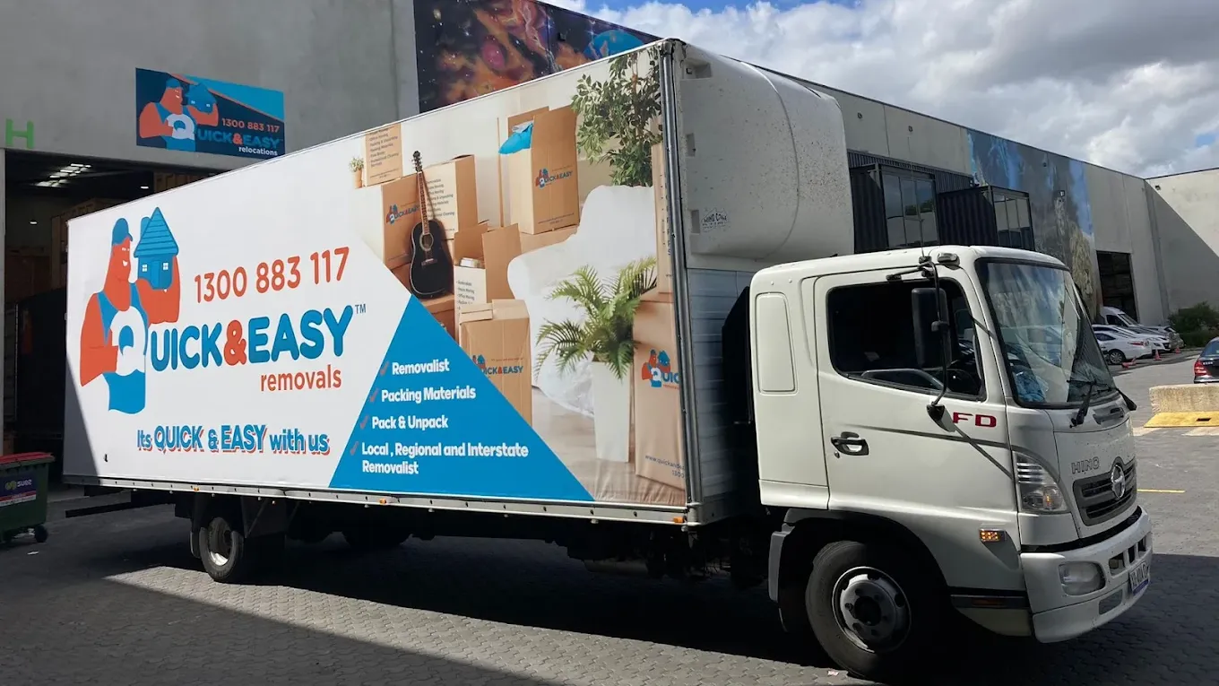A white Hino truck in blue and orange Quick and Easy livery, its side panel reading local, regional and interstate removalist, parked at the roller door of the company depot under a blue sky