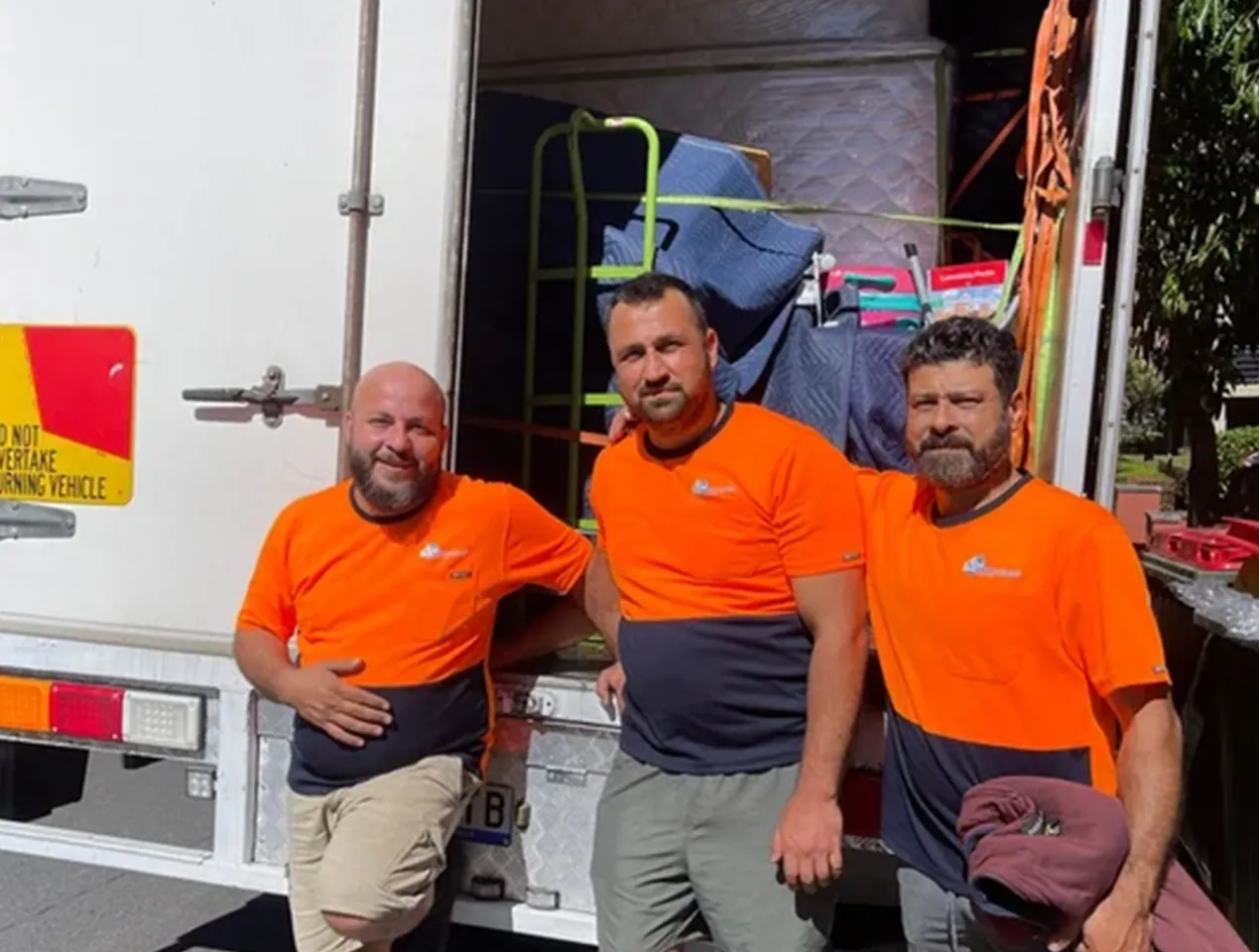 Three Quick and Easy crew members in orange and navy work shirts standing at the open rear of a loaded truck, with a trolley and moving blankets inside