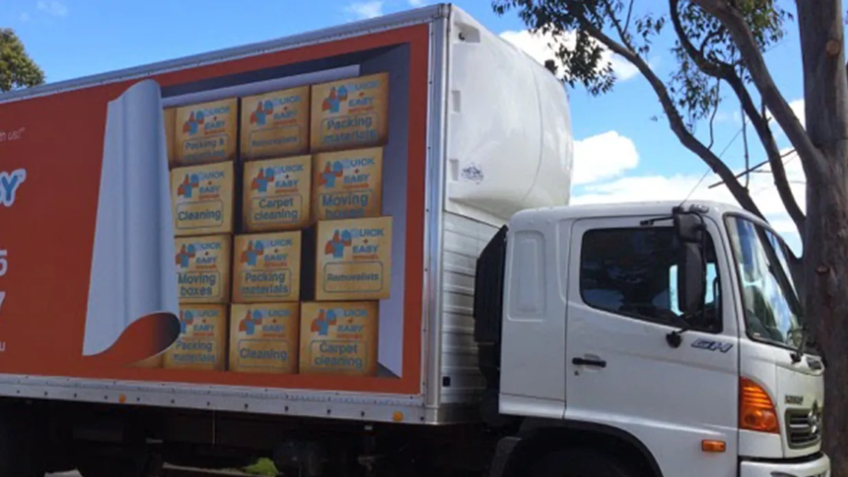 A white Hino truck in orange Quick and Easy livery, its side printed with a wall of carton graphics, parked beside tall gum trees under a blue sky