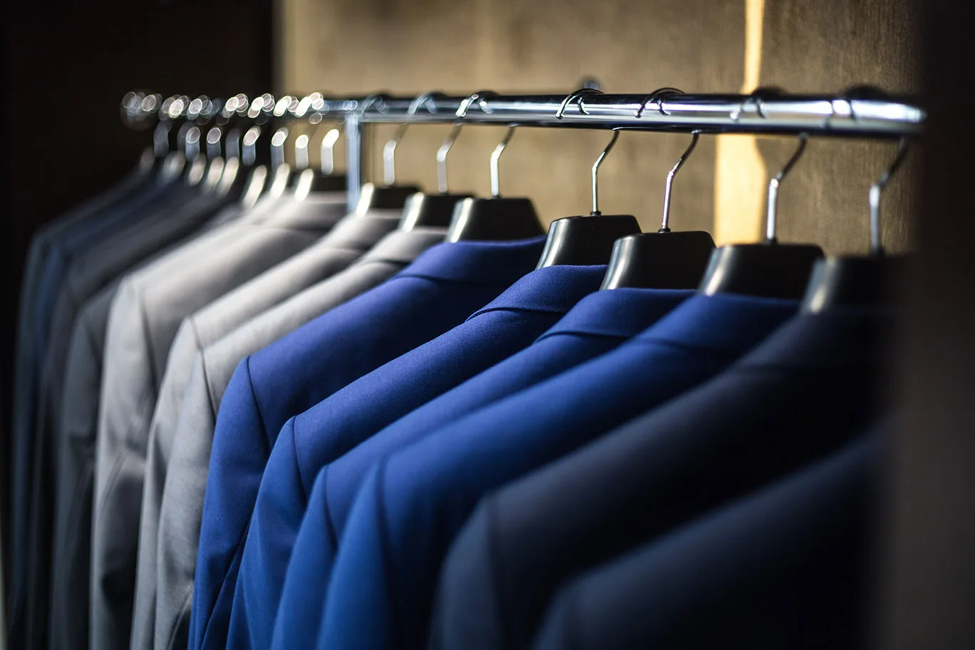 A row of blue, grey and black suit jackets hanging on a rail inside a dark wardrobe