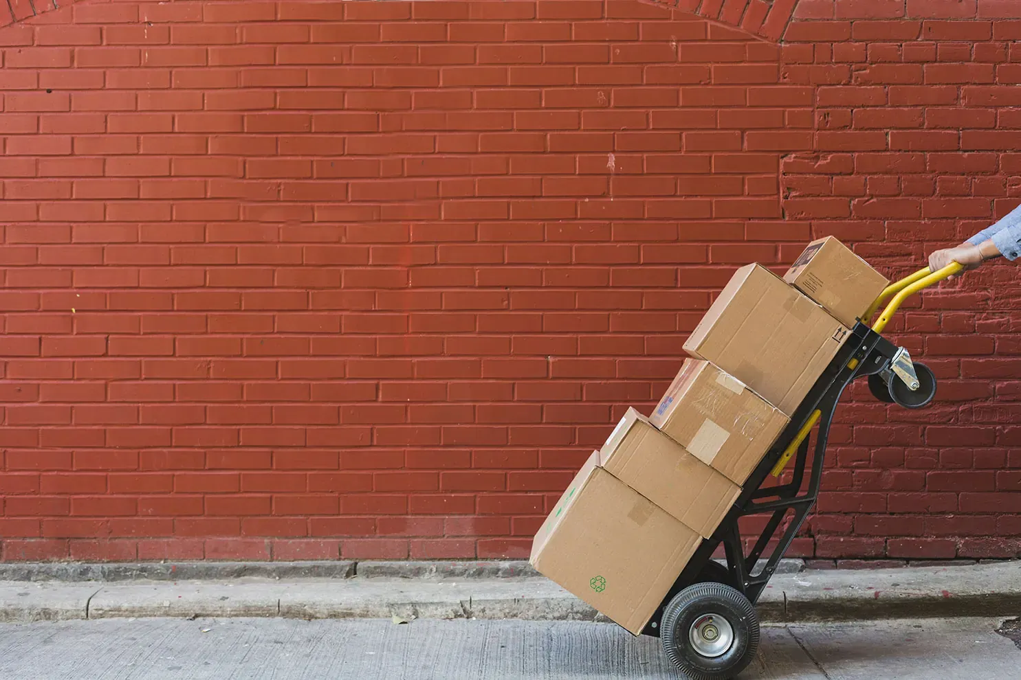 A hand trolley loaded with four cardboard cartons being wheeled along a footpath